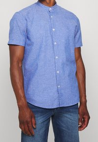 Hombre que lleva una camisa de botones de manga corta, de color azul claro con un pequeño patrón de puntos oscuros, y jeans azules casuales.