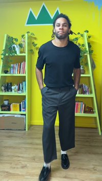 Chemise noire à manches courtes, pantalon gris à larges rayures et jambes larges, chaussures noires, debout dans une pièce colorée avec des étagères vertes et des plantes.