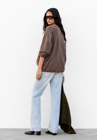 Überdimensionierter brauner Sweatshirt, hellblaue weit geschnittene Jeans, schwarze Stiefeletten mit Absatz, hält einen dunkelgrünen Pullover, vor einem schlichten weißen Hintergrund.