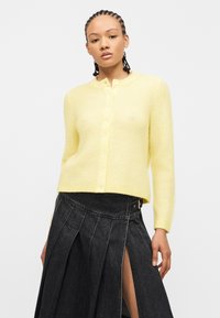 Γυναίκα με πλεγμένα μαλλιά που φοράει κίτρινη ζακέτα με κουμπιά και μαύρη φούστα με πιέτες με ψηλή μέση και σκίσιμο μπροστά.