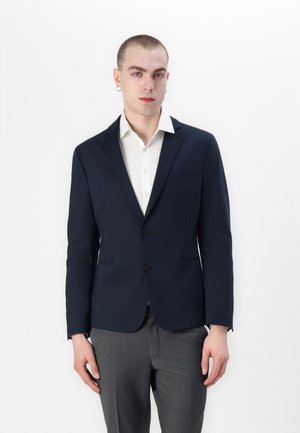 Navy blazer with two buttons, single chest pocket, and sharp lapels. Worn over a white dress shirt. Slim fit and smooth texture.