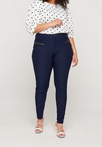 Marineblauwe denim broek met een slim fit, voorzien van gouden ritssluiting aan de voorkant en een klassieke vijfzakken ontwerp. Gepaard met een witte top met stippen.