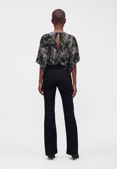 Femme aux cheveux courts portant un chemisier noir à fleurs avec une ouverture dans le dos et un pantalon noir taille haute, debout de dos.