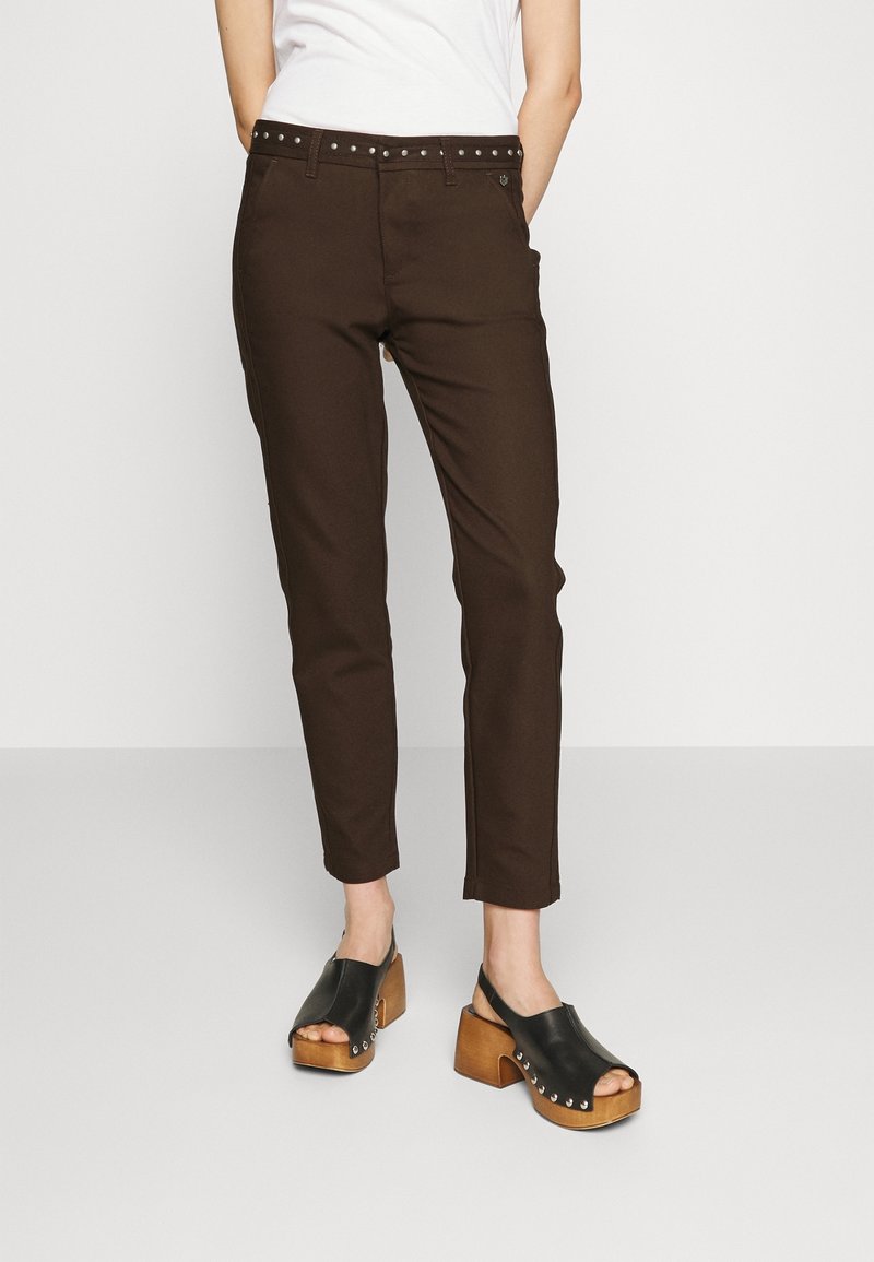 Pantalon marron en mélange de coton, coupe slim, avec une ceinture noire cloutée et des poches latérales. Associé à des sabots en cuir noir à talons en bois.