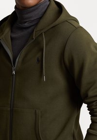 Sudadera con cremallera en verde oliva, con un bolsillo frontal, capucha con cordón y un logo negro en el pecho. Material suave con textura lisa.