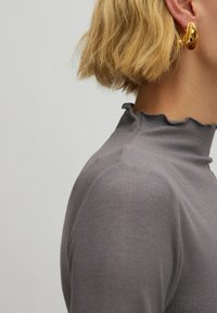 Grauer gerippter Oberteil mit hohem, rüschigem Kragen. Zeigt eine Nahaufnahme der Schulter und des Nackens, mit sichtbaren kurzen, welligen Haaren. Goldenes Ohrring-Detail.