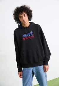 Felpa nera con cappuccio, tasca frontale e testo ricamato colorato in blu e rosso. Indossata sopra jeans azzurri.