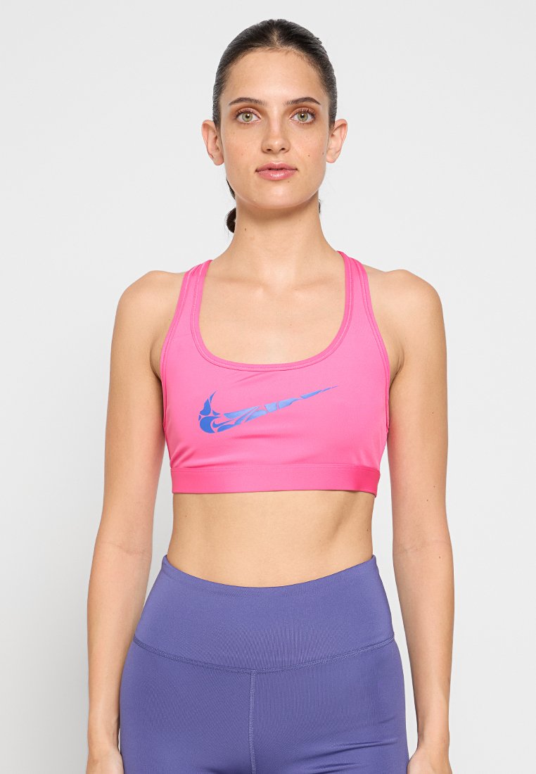 Mujer de pie de frente, vestida con un sujetador deportivo rosa con un logo azul y leggings morados de cintura alta, contra un fondo liso.