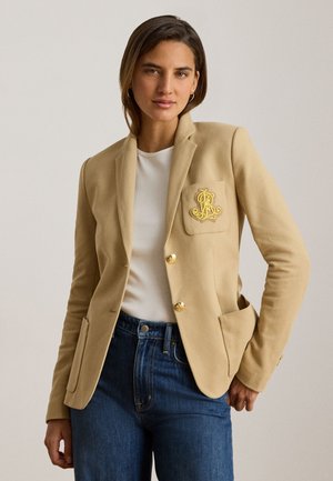 Kobieta ubrana w beżową marynarkę z złotymi guzikami i haftowanym herbem, białą koszulę i niebieskie dżinsy, pozująca na jednolitym jasnym tle.