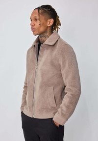 Veste zippée en suédine beige avec col, poches latérales et surface texturée, portée par-dessus une chemise noire, sur un fond uni.