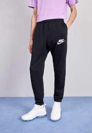 Zwarte Nike trainingsbroek met een elastische tailleband, met een wit en paarse logo op het linkerbeen, gecombineerd met witte sportschoenen.