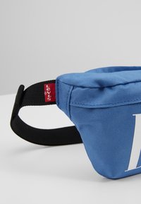 Sac banane en tissu bleu avec sangle noire réglable et étiquette rouge visible du logo Levi's à l’extrémité de la sangle.