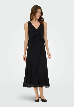 Femme portant une robe midi noire sans manches avec un col en V volanté et des chaussures slingback noires à bouts pointus, debout devant un fond uni.