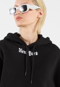 Felpa nera con cappuccio che presenta "New York" in carattere gotico bianco, abbinata a occhiali da sole bianchi e angolari, evidenziando una texture liscia e un design minimalista.
