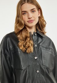 Jachetă din piele neagră, cu nasturi, două buzunare pe piept și guler ascuțit. Textură netedă și detalii elegante în cusături.