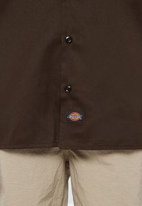 Brown buttoned shirt with Dickies logo above beige textured pants waistline.