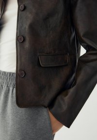 Barna bőrkabát négyzetmintával, két gombbal, mellső zsebbel és sima textúrával. Szürke elasztikus derékpántos nadrággal párosítva.