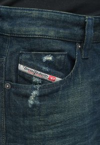 Kék farmernadrág kopott, elöregedett részletekkel, a kis elülső zsebben látható a Diesel Industry Denim Division címke.