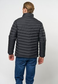 Črna puffer jakna z vodoravnim pre�šitim vzorcem, visokim ovratnikom in dolgimi rokavi, v kombinaciji z modrimi kavbojkami. Gladka tekstura in lahka zasnova.