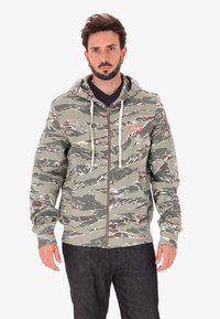 Uomo che indossa una felpa con cappuccio a zip nei colori verde e marrone mimetico, con lacci bianchi e pantaloni scuri, in piedi su uno sfondo bianco.