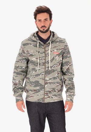 Man in een groen en bruin camouflage zip-up hoodie met witte trekkoorden en donkere broek, staande tegen een witte achtergrond.