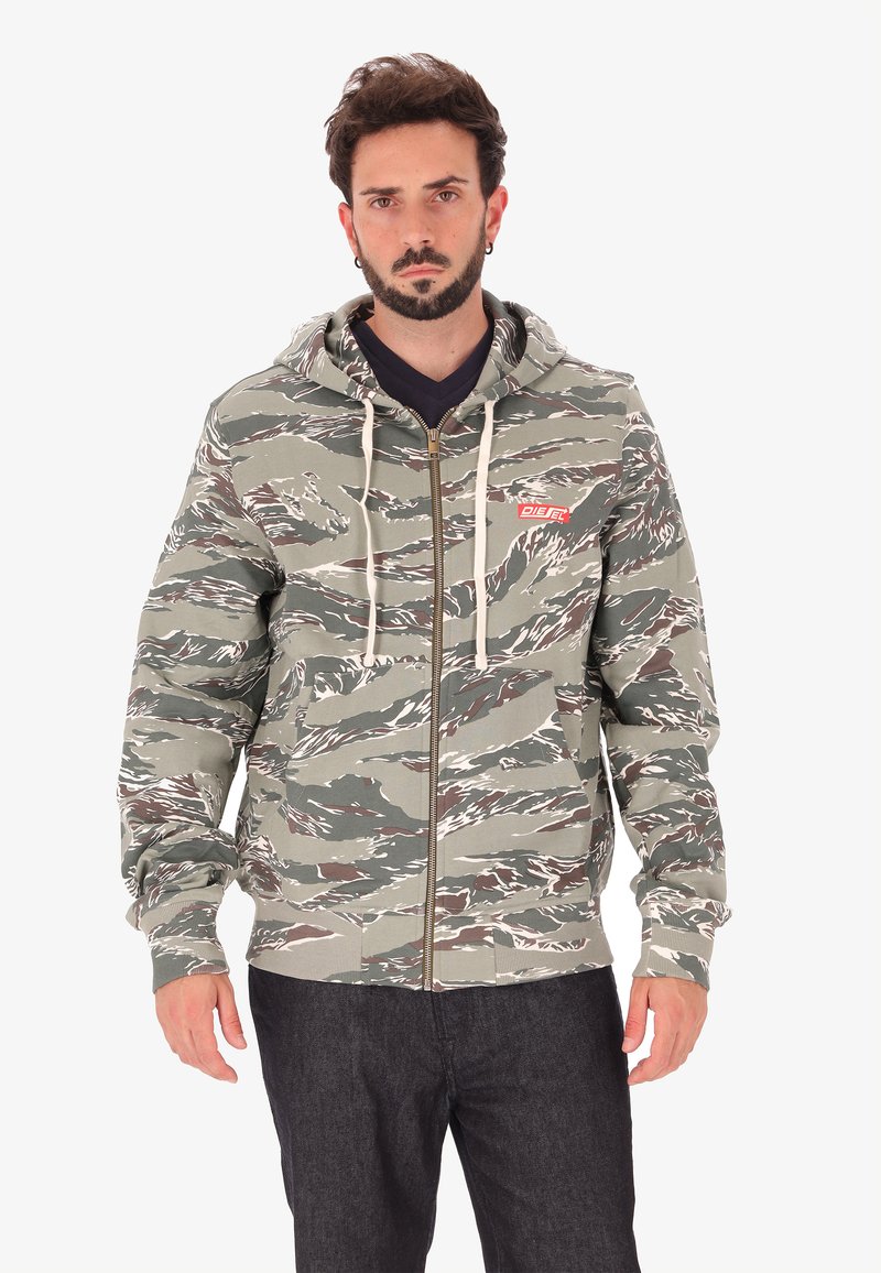 Uomo che indossa una felpa con cappuccio a zip nei colori verde e marrone mimetico, con lacci bianchi e pantaloni scuri, in piedi su uno sfondo bianco.