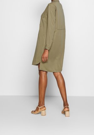 Osoba ubrana w luźną sukienkę koszulową w kolorze oliwkowozielonym z długimi rękawami oraz beżowe sandały na słupku, stojąca na tle białego tła.