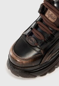 Tênis de couro preto com detalhes em castanho, sola de borracha texturizada, bico redondo e etiqueta de logo na língua. Inclui atacadores castanhos e colarinho acolchoado.