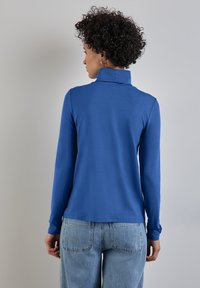 Blaues Langarmoberteil mit hohem Kragen, aus glattem Stoff gefertigt. Besitzt einen lässigen Schnitt und einen geraden Saum. Kombiniert mit hellen Jeans.