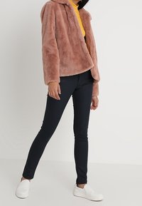 Rosa fuskpälsjacka med huva, med en avslappnad passform. Bärs över en senapsgul topp och tillsammans med marinblå skinnybyxor och vita slip-on skor.