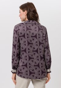 Blouse à manches longues en violet foncé avec un motif géométrique blanc. Dotée d'un col et de poignets accentués avec des rayures. Texture de tissu douce.