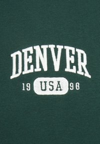 La tela de algodón verde presenta texto bordado en blanco: "DENVER" arqueado sobre "USA" y "19 98" debajo, con una textura suave.