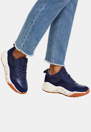 Persoon draagt blauwe en marineblauwe chunky sneakers met oranje zolen en lichtblauwe jeans met rafelige zoom tegen een witte achtergrond.
