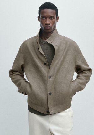 Uomo che indossa una giacca kaki con bottoni sopra un maglione oliva e pantaloni bianchi, in piedi con le mani nelle tasche contro uno sfondo semplice.