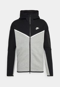 Sudadera con cremallera Nike en negro y gris, con capucha, logo blanco en el pecho, banda en chevrón blanca y bolsillo con cremallera en la manga izquierda.