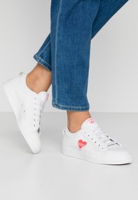 Vita canvas sneakers har en röd hjärtformad design och rosa detaljer på tungan. Sulan är vit och snörena är standard. Undviker komplexa mönster.