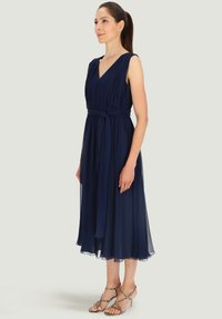 Granatowa sukienka do kolan z dekoltem w serek, marszczonym materiałem i związaną talią, wykonana z lekkiego materiału; zestawiona z wzorzystymi sandałami na paskach.