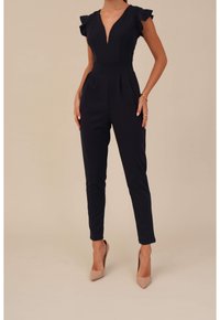 Mörkblå overall med volangdetaljer vid axlarna, djup V-ringning, avsmalnande ben och veckad front. Bärs med nude högklackade skor.