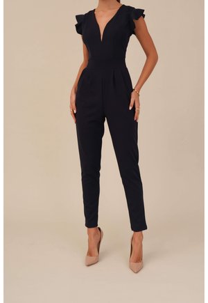 Mörkblå overall med volangdetaljer vid axlarna, djup V-ringning, avsmalnande ben och veckad front. Bärs med nude högklackade skor.