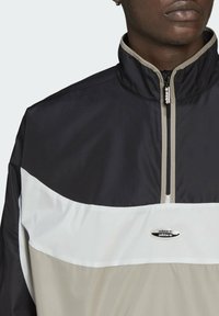 Man som bär en svart, vit och beige Adidas zip-up jacka med logotyp på bröstet och dragkedja, visad från axlarna till läpparna.