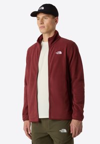 Giacca in pile borgogna con colletto alto e zip completa, con un logo bianco sul lato sinistro del petto. Indossata sopra una camicia grigia chiara.