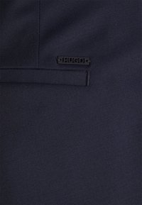 Tissu bleu marine foncé avec une petite étiquette noire rectangulaire marquée « HUGO », fixée près d'une couture horizontale du tissu.