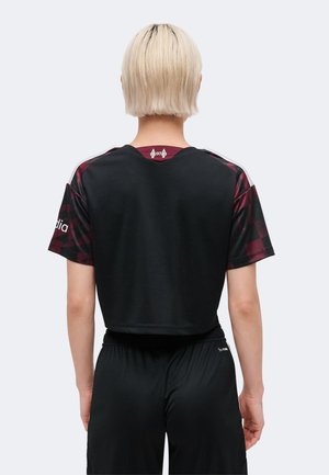 Persona con cabello rubio corto que lleva una camiseta deportiva de manga corta negra y burdeos y pantalones cortos negros, de espaldas a la cámara.