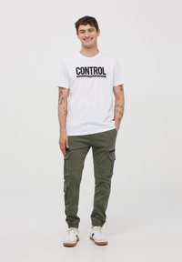 Biała koszulka z krótkim rękawem z napisem "CONTROL" wyboldowanym czarnym kolorem, zestawiona z oliwkowymi spodniami cargo z wieloma kieszeniami i elastycznymi mankietami.