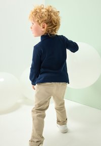 Marineblauwe fleece trui met een hoge kraag, gecombineerd met beige katoenen broek en witte sneakers. Lichte achtergrond met witte ballonnen.
