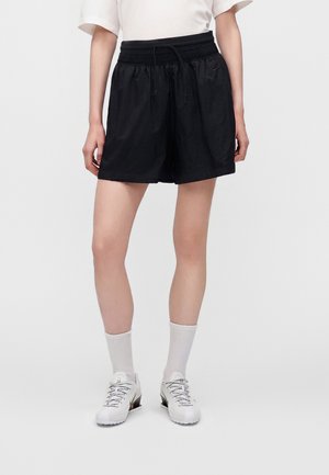 Person iført sorte løse shorts med snøre, hvide sokker, hvide sneakers og en kortærmet hvid skjorte, står på en ensfarvet baggrund.