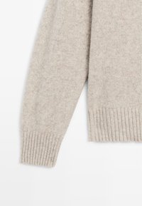 Beżowy sweter z dzianiny z prążkowanymi mankietami i obrębem. Miękka tekstura, lekko zróżnicowany kolor z drobnymi plamkami. Zawiera boczne rozcięcie.