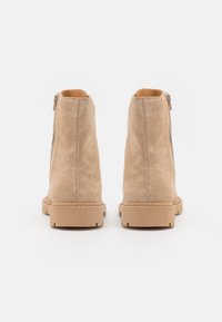 Un paio di stivaletti in suede beige alla caviglia con cerniere laterali, visti da dietro, su uno sfondo bianco.