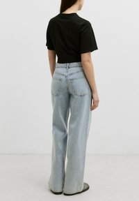 Mujer de pie de espaldas, vistiendo una camiseta negra de manga corta, unos jeans de tiro alto y pierna ancha en azul claro, y zapatos grises, frente a un fondo liso.