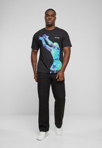 Czarny bawełniany t-shirt z kolorową grafiką w stylu termicznym przedstawiającą postać sięgającą w górę. W połączeniu z czarnymi spodniami i białymi trampkami.